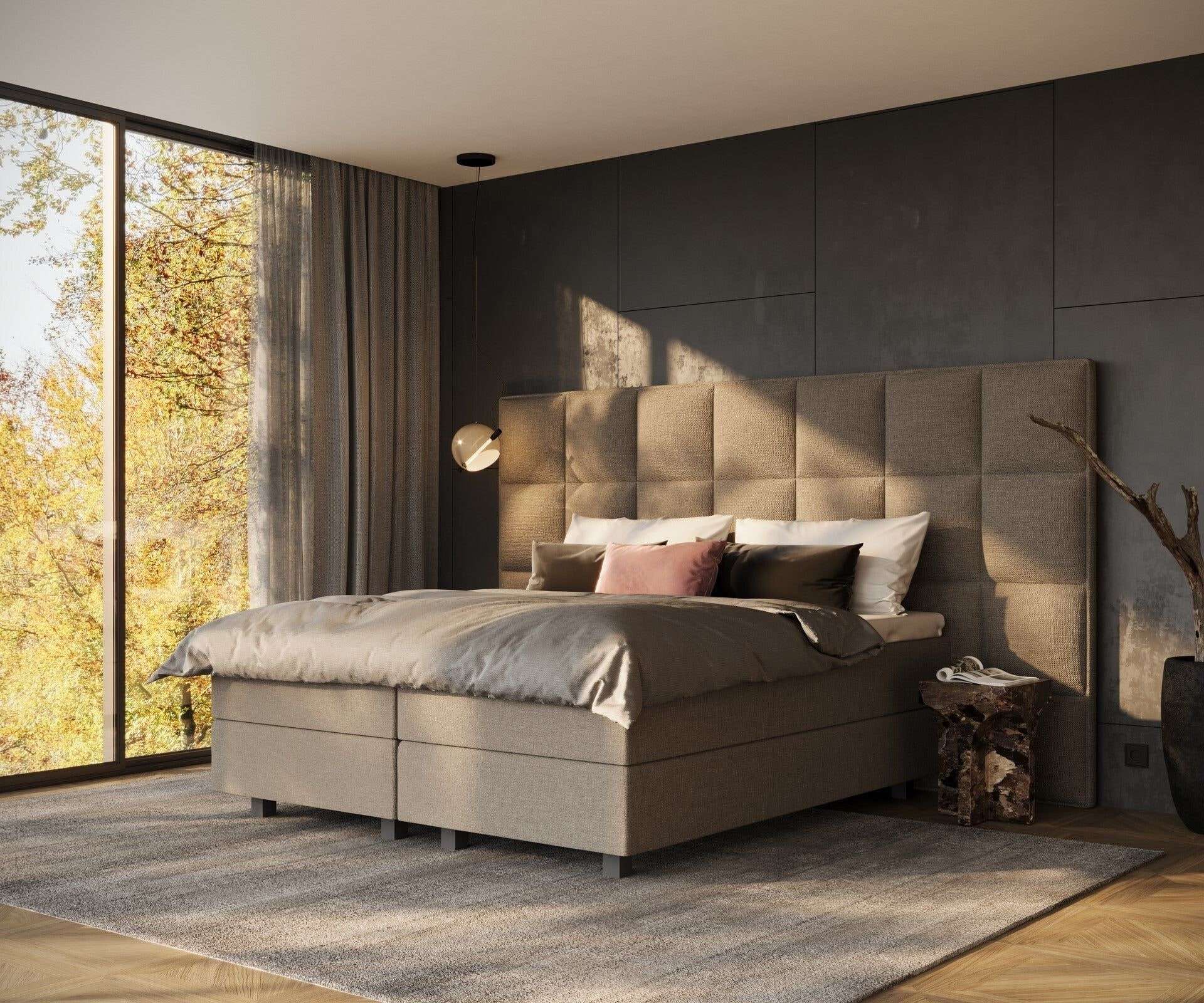 Een brede boxspring met een hoog gestoffeerd hoofdbord tegen een donkere wand, in laag avondlicht